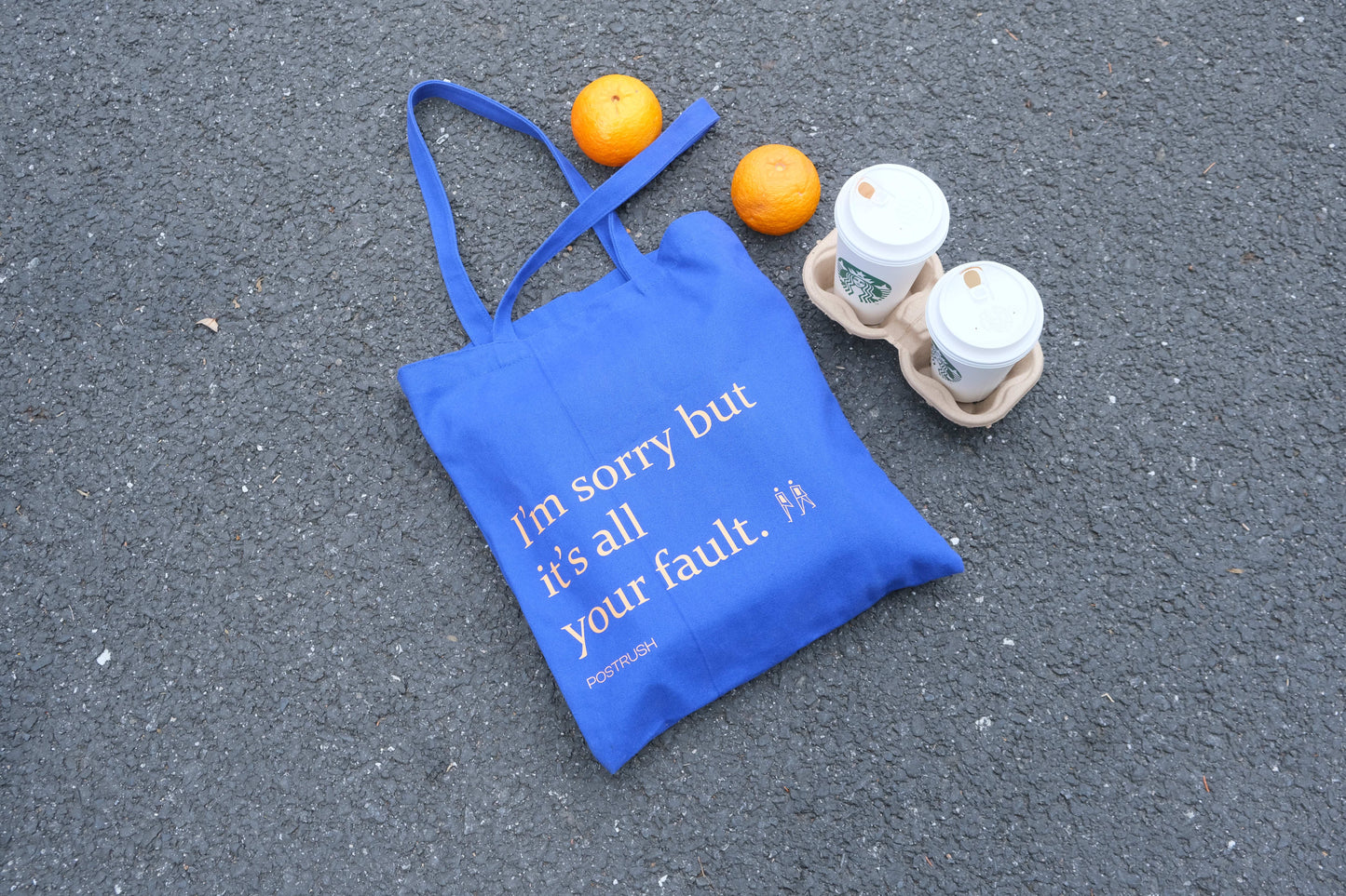 Blue canvas tote bag with the text ‘I’m sorry but it’s all your fault,’ placed on asphalt next to takeaway coffee cups and oranges.