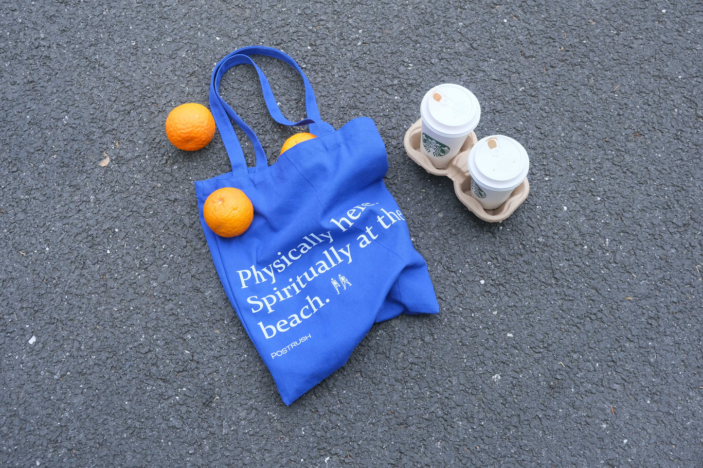 Blue canvas tote bag with the text ‘Physically here. Spiritually at the beach,’ placed on asphalt with oranges and takeaway coffee cups.