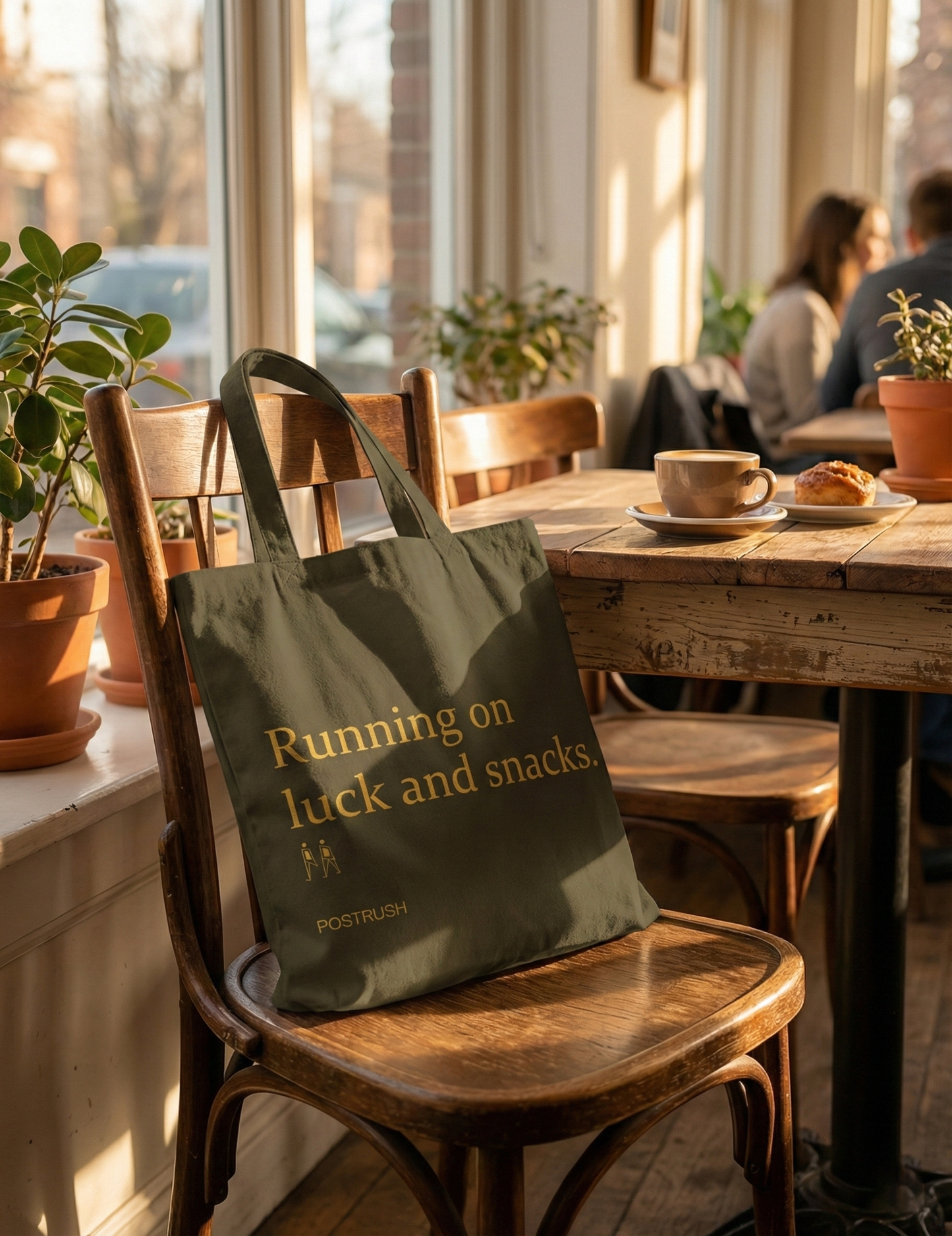 Brown canvas tote bag with the quote “Running on luck and snacks.” placed on a wooden chair in a sunlit café.
