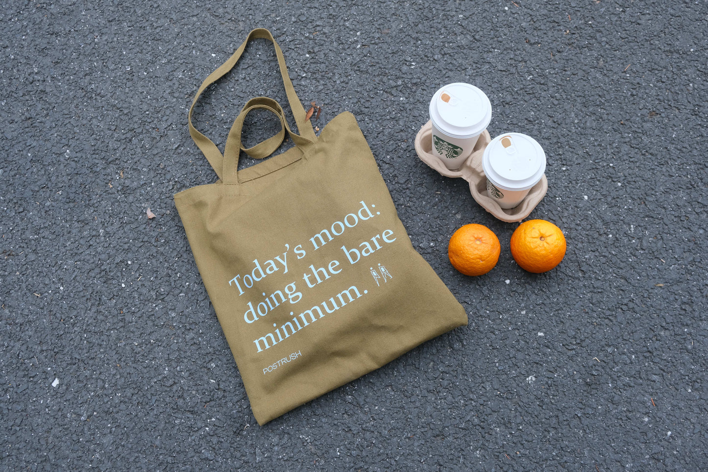 Brown canvas tote bag with the text ‘Today’s mood: doing the bare minimum,’ placed on asphalt next to two takeaway coffee cups and oranges.