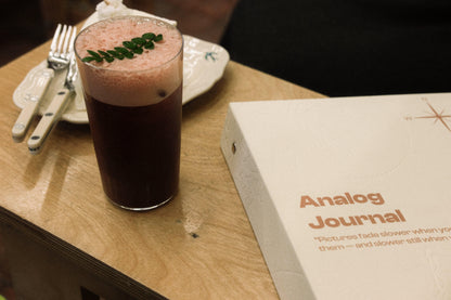 Glass of dark liquid with a pink top on a wooden table next to a 'Analog Journal' album.