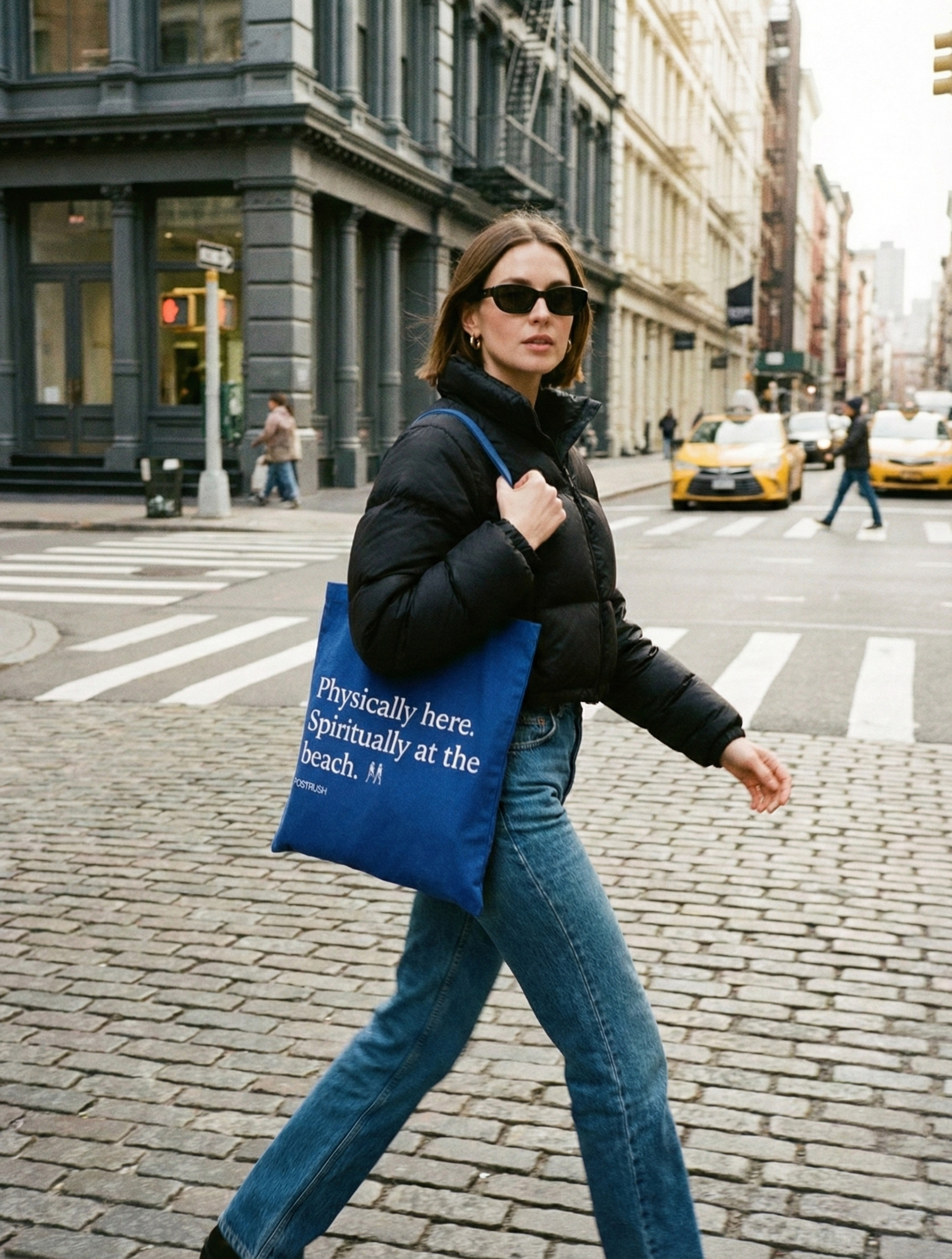 Woman crossing a city street carrying a blue canvas tote bag with the text ‘Physically here. Spiritually at the beach.’