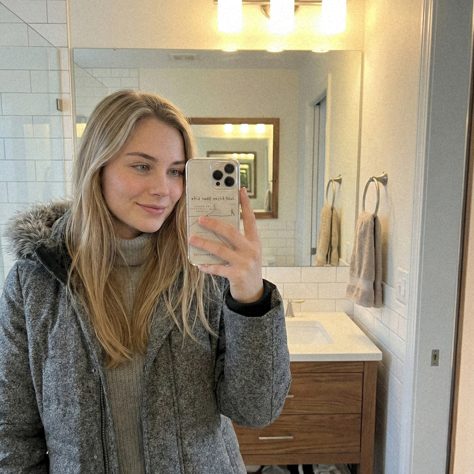 Person taking a bathroom mirror selfie while holding a clear phone case with “Just Enjoy Your Life” text and minimalist line art design.