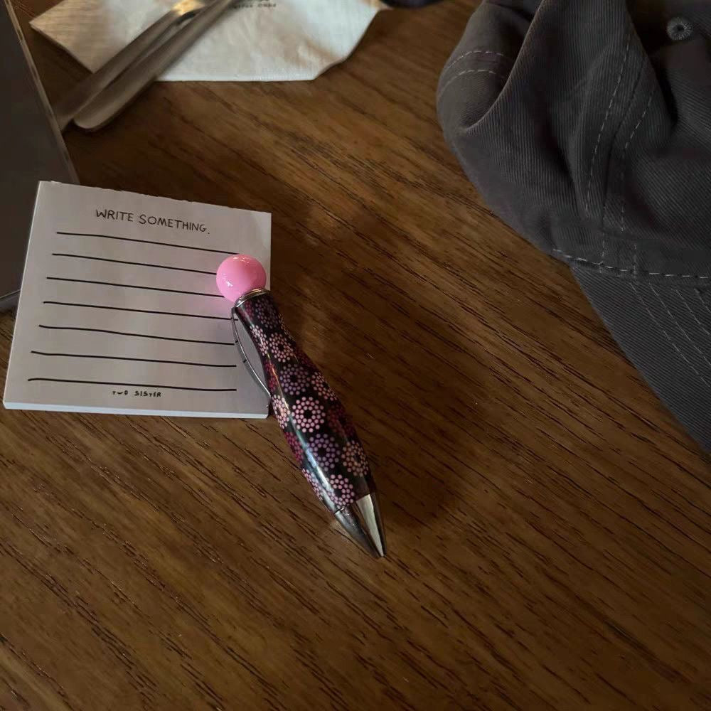 Pink polka dot ballpoint pen resting on a wooden table next to a small paper note.