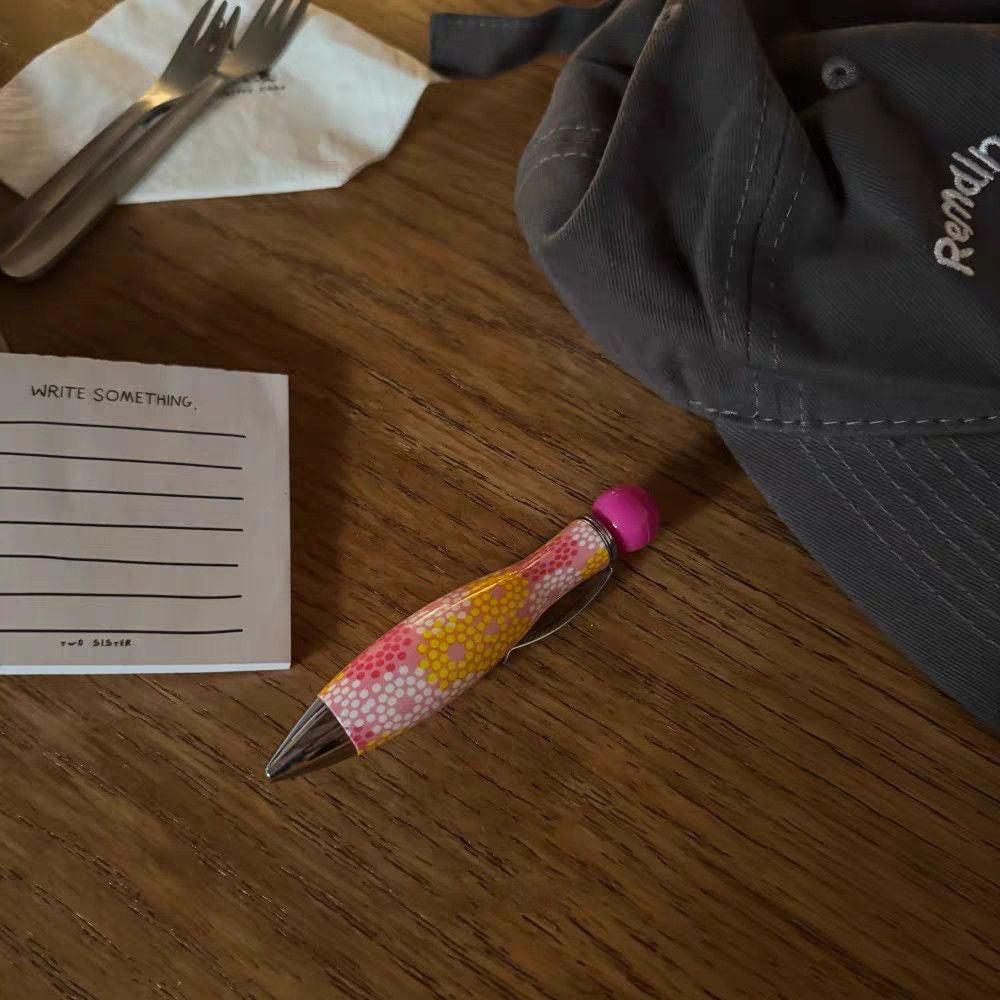 Pink and yellow polka dot ballpoint pen resting on a wooden table next to a paper note.