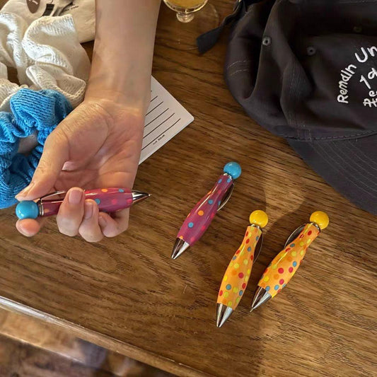 Hand holding a pink polka dot ballpoint pen with other polka dot pens in multiple colors displayed on a wooden table.