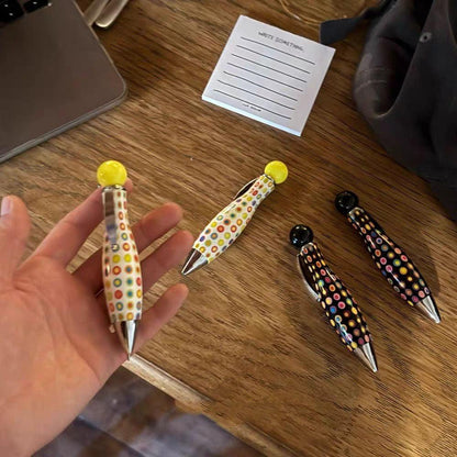 Multiple polka dot ballpoint pens in different colorways displayed on a wooden table, with one held in hand.