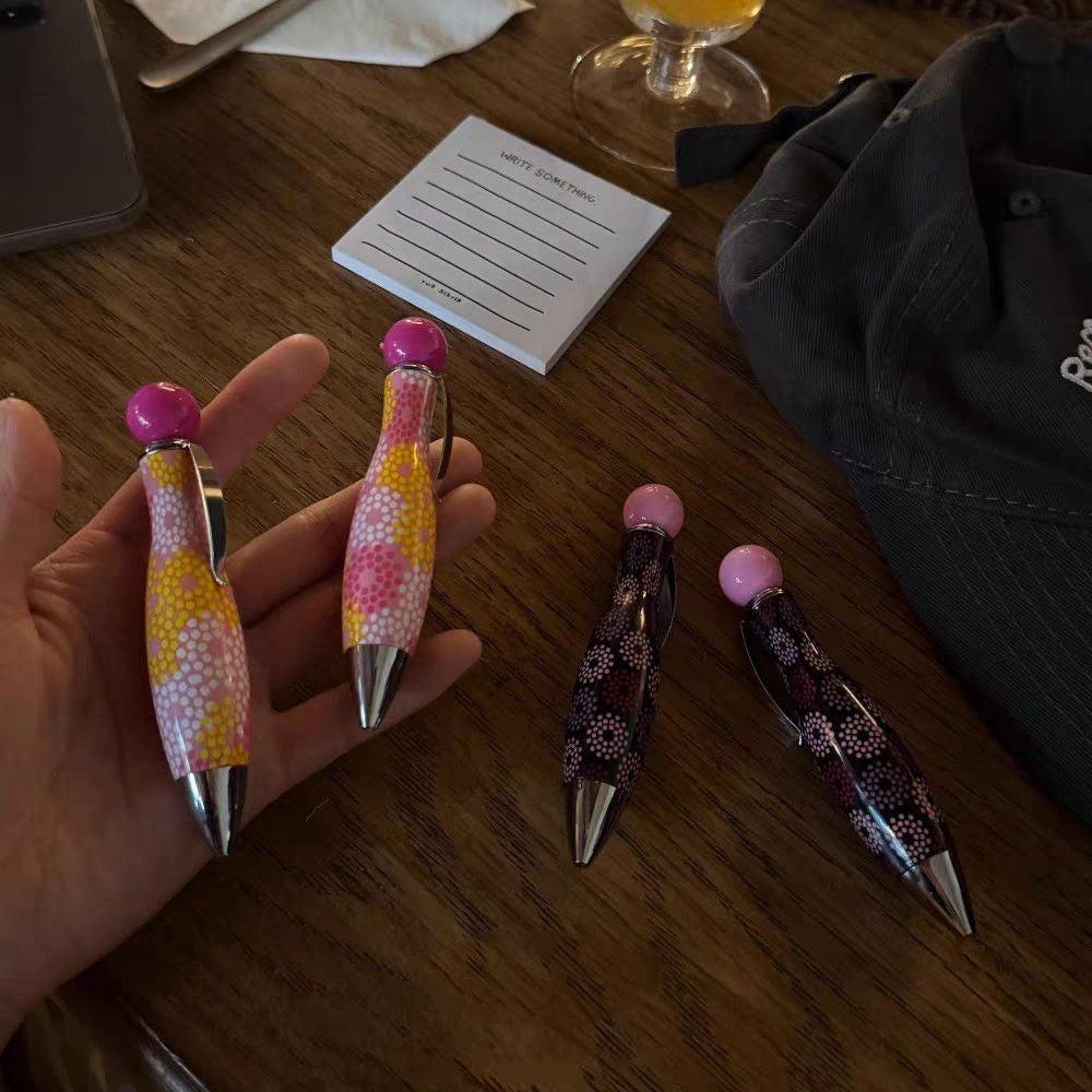 Hand holding two polka dot ballpoint pens with additional pink and black patterned pens displayed on a wooden table.