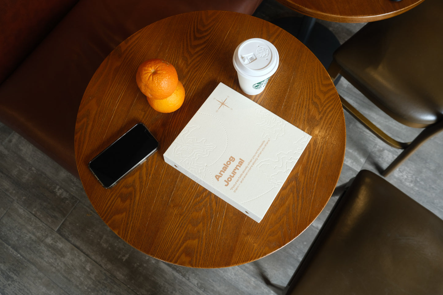 A white PostRush analog journal and scrapbook album on a wooden cafe table next to a coffee cup, two oranges, and a smartphone.