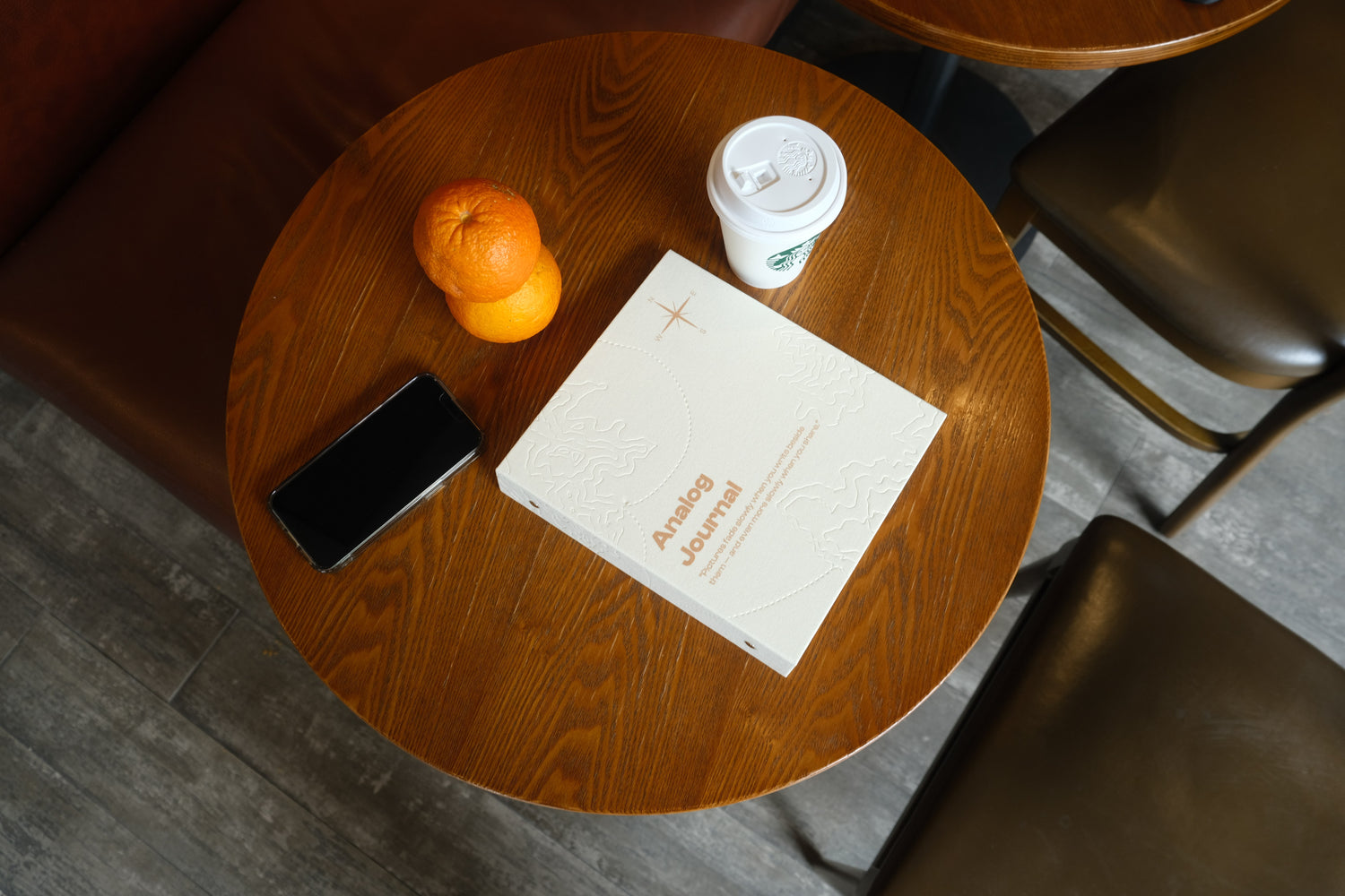 A white PostRush analog journal and scrapbook album on a wooden cafe table next to a coffee cup, two oranges, and a smartphone.