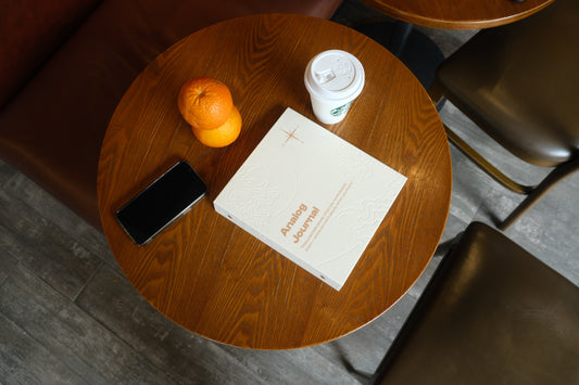 A white PostRush analog journal and scrapbook album on a wooden cafe table next to a coffee cup, two oranges, and a smartphone.