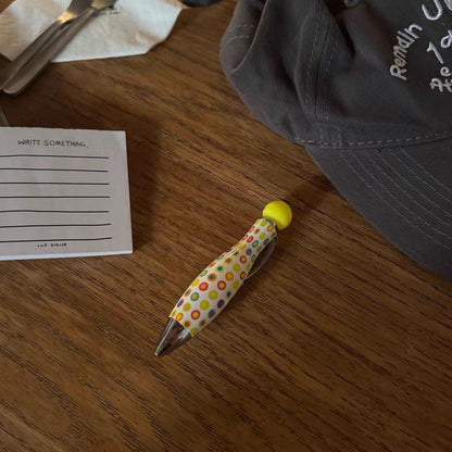 Yellow polka dot ballpoint pen resting on a wooden table next to a paper note and a fabric bag.